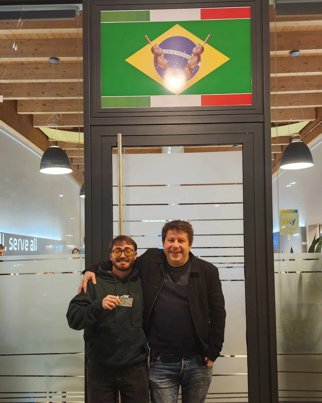 Dos clientes de Speranza posando frente a un cartel con las banderas de Brasil e Italia